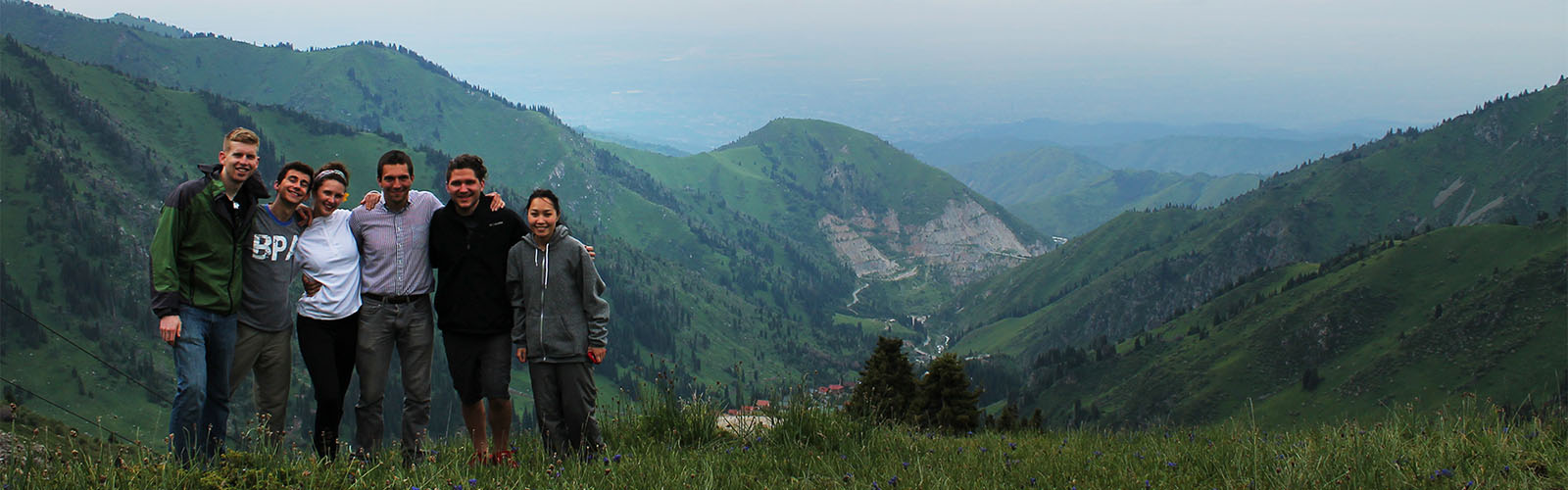 Six students stand on a grassy mountain, and there are many other grassy mountains in the background. 